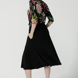 A size 10 woman wears the Jules top in Bloomsbury print. A curved neckline style with 3/4 sleeves. Made in easy care jersey fabric with a beautiful floral print with red, pink, orange, green and vanilla on a black base. Made in Australia for women size 8-24. Styled back with the Berit skirt in black.