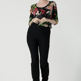 A size 10 woman wears the Jules top in Bloomsbury print. A curved neckline style with 3/4 sleeves. Made in easy care jersey fabric with a beautiful floral print with red, pink, orange, green and vanilla on a black base. Made in Australia for women size 8-24. Styled back with the slim fit Brooklyn pant in black.