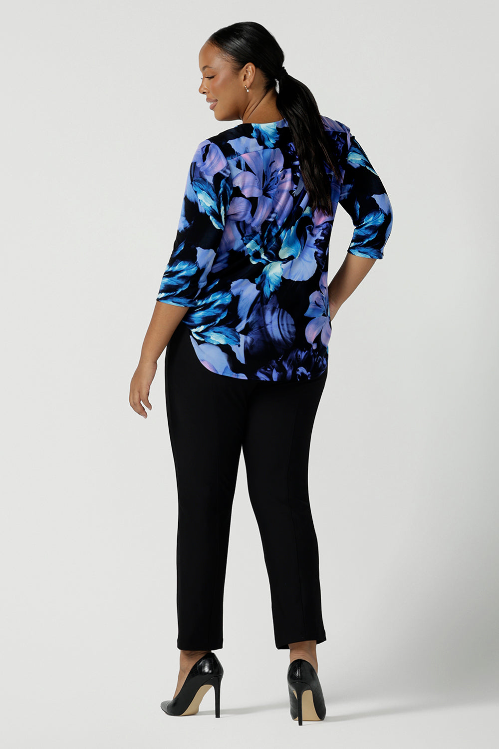 Back view of a size 16 woman wears the Jules top in Blue Lily. A Scoop neck style we a 3/4 sleeve. Digitally printed with a blue Lily Print. Size inclusive fashion, made in Australia for women size 8 - 24. Styled back with Black slim leg brooklyn pants.