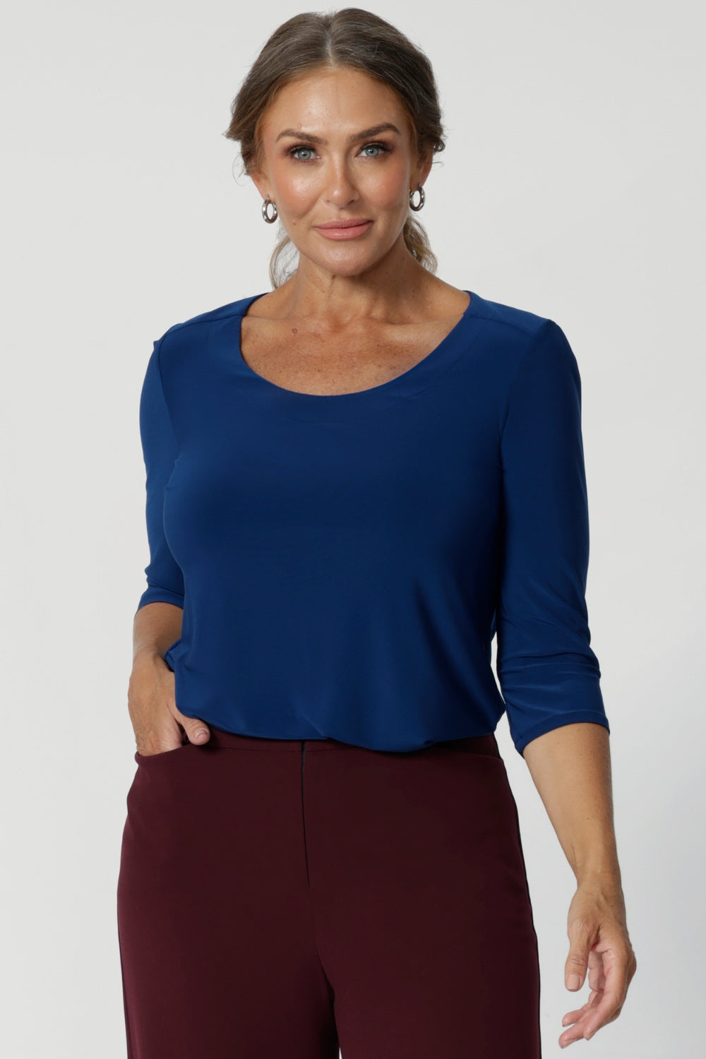 The Jules Top is the ultimate collarless tailored shirt, designed for both workwear and weekend wear. Crafted from a deep blue dry-touch jersey fabric, round neck top offers a soft stretch that moves with you, ensuring all-day comfort without compromising on style. Size 12 woman pairs her Jules top with wine coloured workwear trousers. 