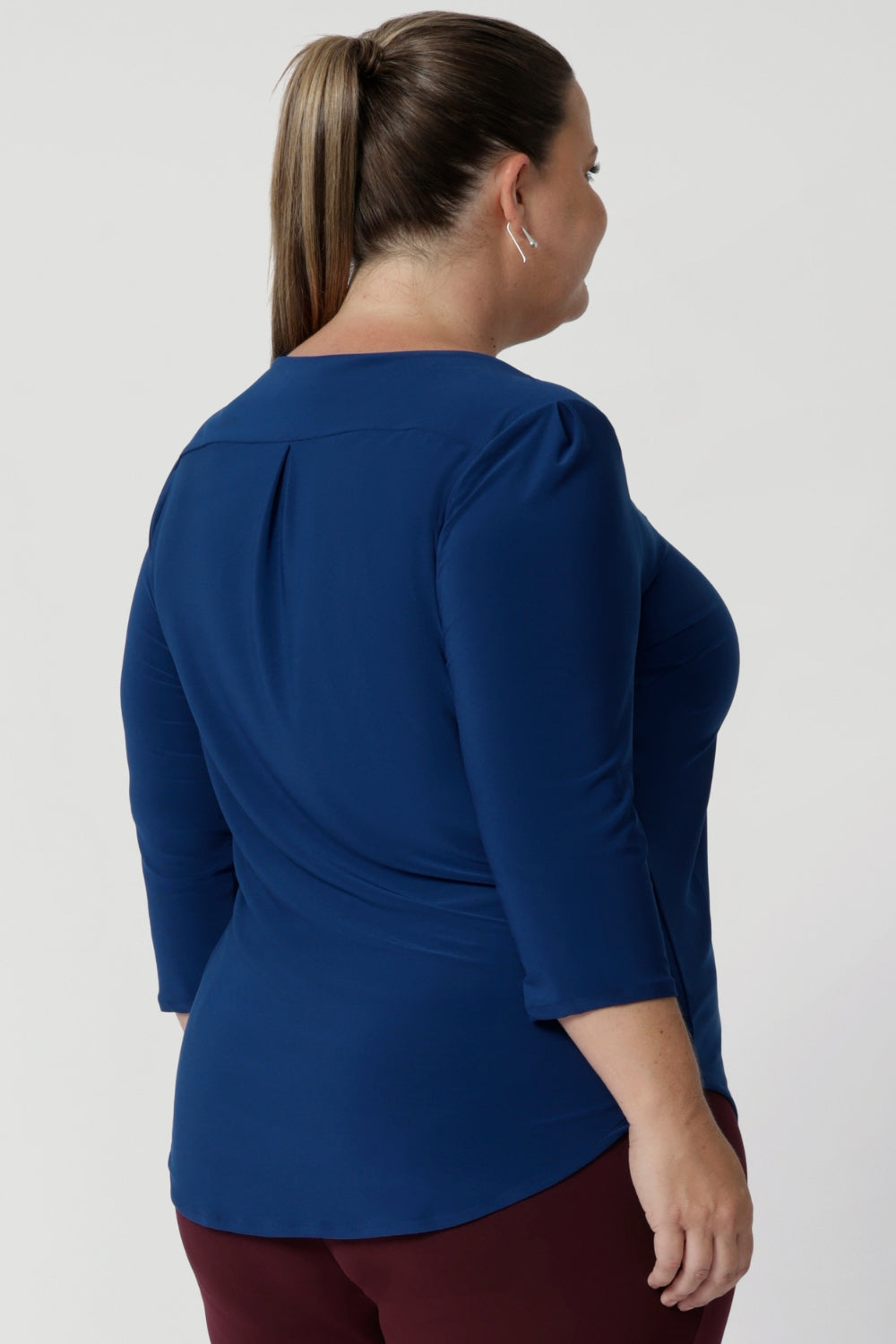 Back view image of plus size woman shows her wearing deep blue workwear top showcasing back shoulder yoke with a central pleat below that provides extra shape and dimension, enhancing the top’s tailored feel. Available in sizes 8-24. 