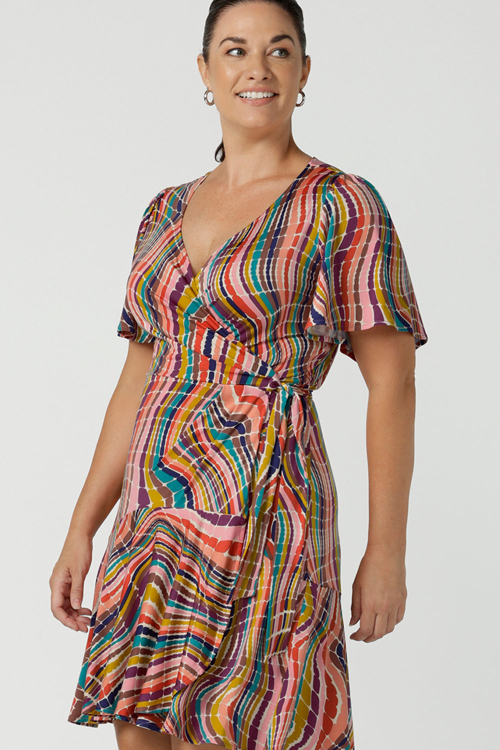 Close up of a women in a size 12 wears the Julie wrap dress in Kaleidoscope rainbow stripe. With a tier frill hem and flutter sleeves, this dress is flirty and great for the party season. Made in Australia size 8 - 24.