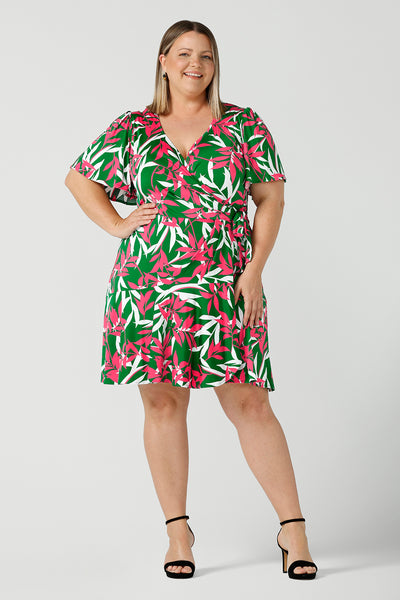 Curvy happy woman wears a size 18 wrap dress with frill hemline, flutter sleeves and wrap ties. Designed in a soft jersey material and featuring a green, fuchsia and white leaf print. Made in Australia for women size 8 - 24. 