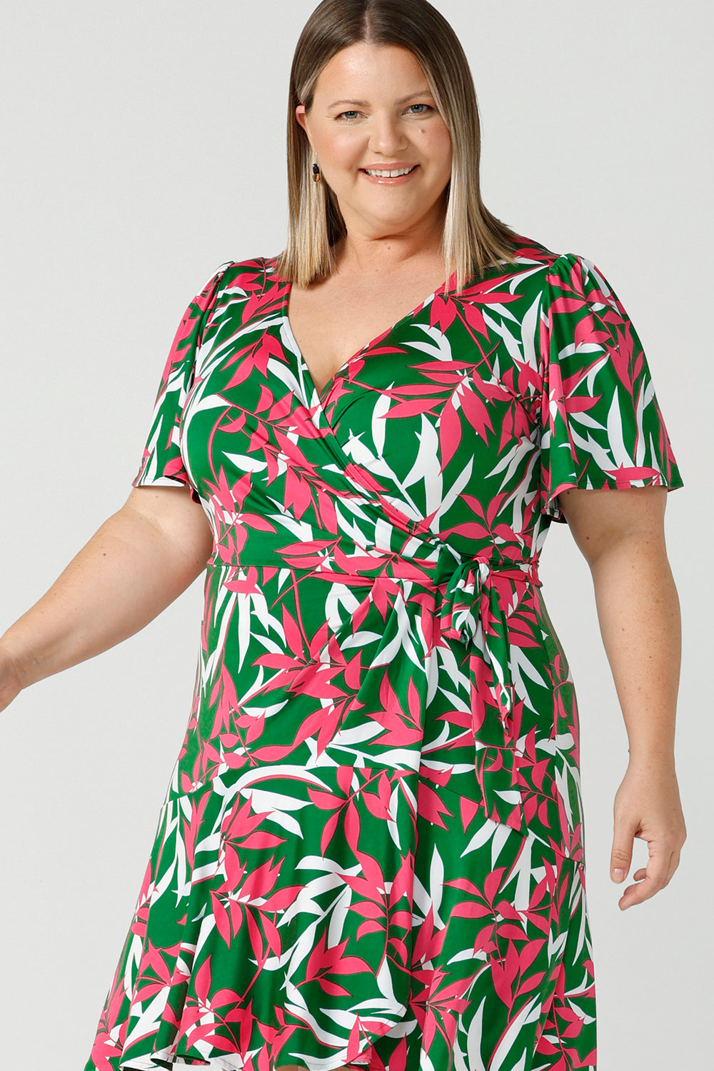 Close up details of a curvy happy woman wears a size 18 wrap dress with frill hemline, flutter sleeves and wrap ties. Designed in a soft jersey material and featuring a green, fuchsia and white leaf print. Made in Australia for women size 8 - 24.