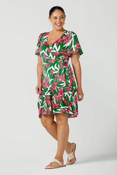A smiley woman in a size 12 jersey dress wears the Julie Dress that is a petite height friendly wrap dress. Designed in a green base fuchsia printed leaf print. With flutter sleeves and a functioning wrap tie with a frill hem. Designed and made in Australia for women size 8 - 24. 