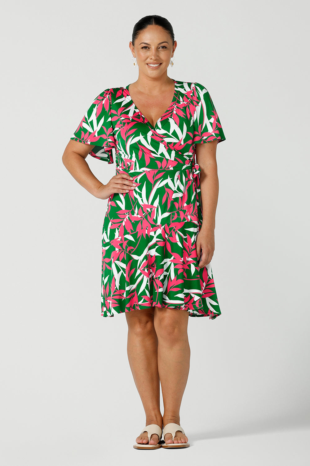 A smiley woman in a size 12 jersey dress wears the Julie Dress that is a petite height friendly wrap dress. Designed in a green base fuchsia printed leaf print. With flutter sleeves and a functioning wrap tie with a frill hem. Designed and made in Australia for women size 8 - 24.