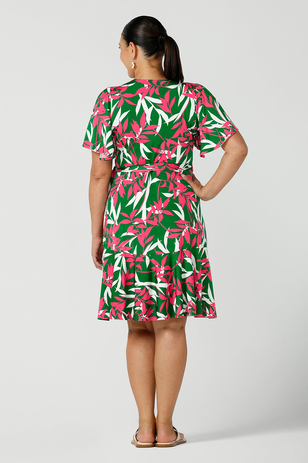 Back view of a woman in a size 12 jersey dress wears the Julie Dress that is a petite height friendly wrap dress. Designed in a green base fuchsia printed leaf print. With flutter sleeves and a functioning wrap tie with a frill hem. Designed and made in Australia for women size 8 - 24.