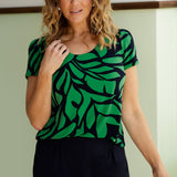 A size 10 woman wears the Sawyer Top in Green Jungle. A scoop neckline with a short sleeve. Soft jersey fabric for easy care. Styled back with Black Indi pants. Made in Australia for women size 8-24.