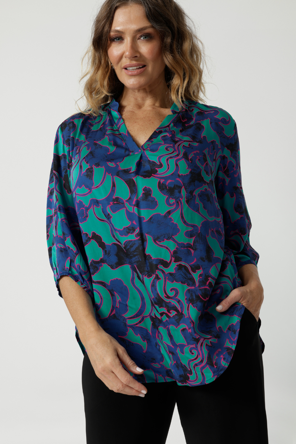 Close up image of an over 40 woman wearing a V neck, light weight shirt with an abstract coloured designed fabric. The fabric colours display, aqua, navy and pink. A leisure workwear shirt for women made in Australia by Australian and New Zealand women. Leina & Fleur is women's clothing label, available sizes include 8 to 24. Shop this comfortable work top in plus size and petite sizes.