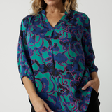 Close up image of an over 40 woman wearing a V neck, light weight shirt with an abstract coloured designed fabric. The fabric colours display, aqua, navy and pink. A leisure workwear shirt for women made in Australia by Australian and New Zealand women. Leina & Fleur is women's clothing label, available sizes include 8 to 24. Shop this comfortable work top in plus size and petite sizes.