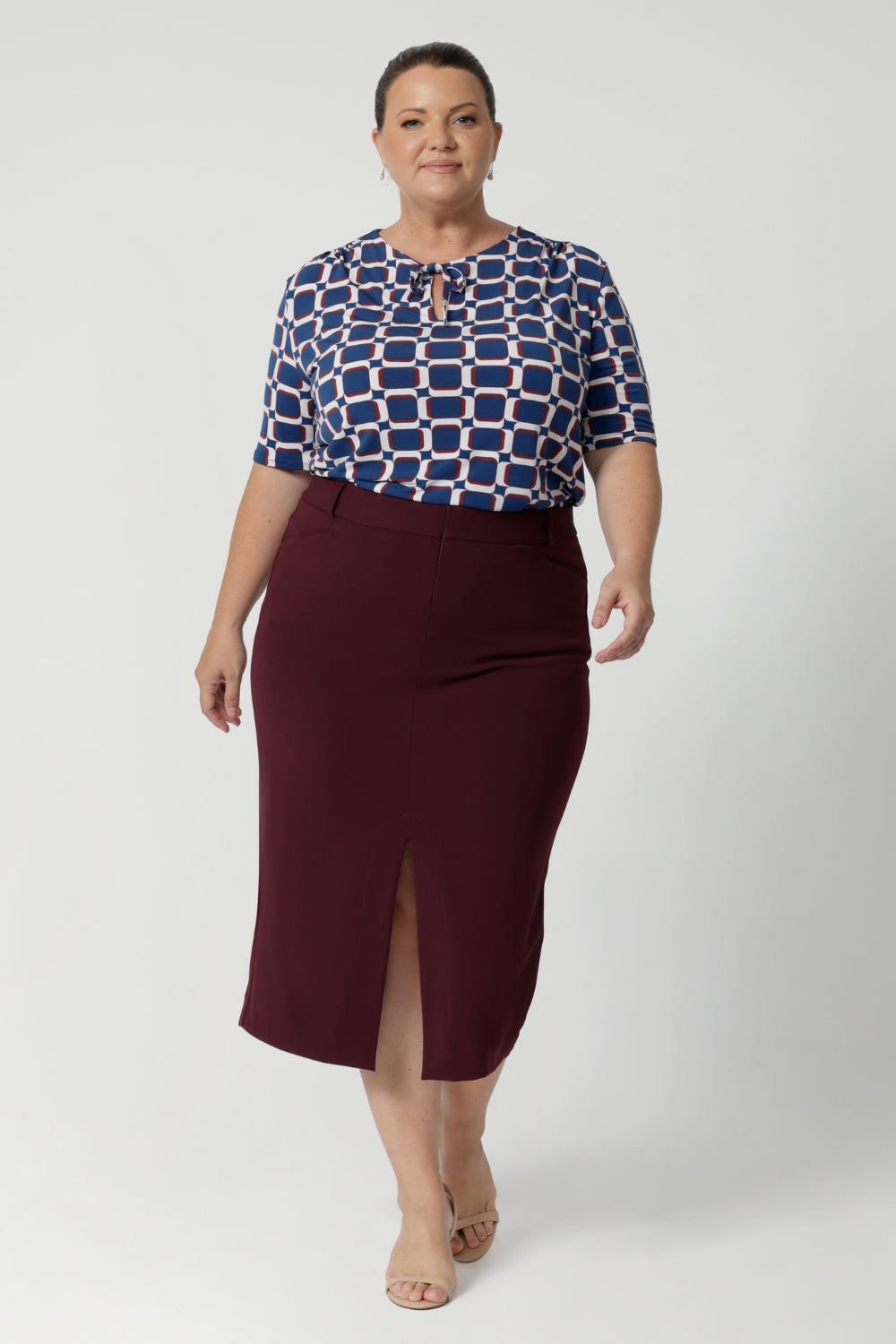 Featuring elbow-length sleeves, this top provides just the right amount of coverage while maintaining a relaxed and chic look. The stitching detail around the front-facing, complemented by thin fabric neckties, adds a tailored touch, perfect for elevating your everyday outfits. Plus size woman wears tie neck top with wine coloured midi skirt and nude heels.