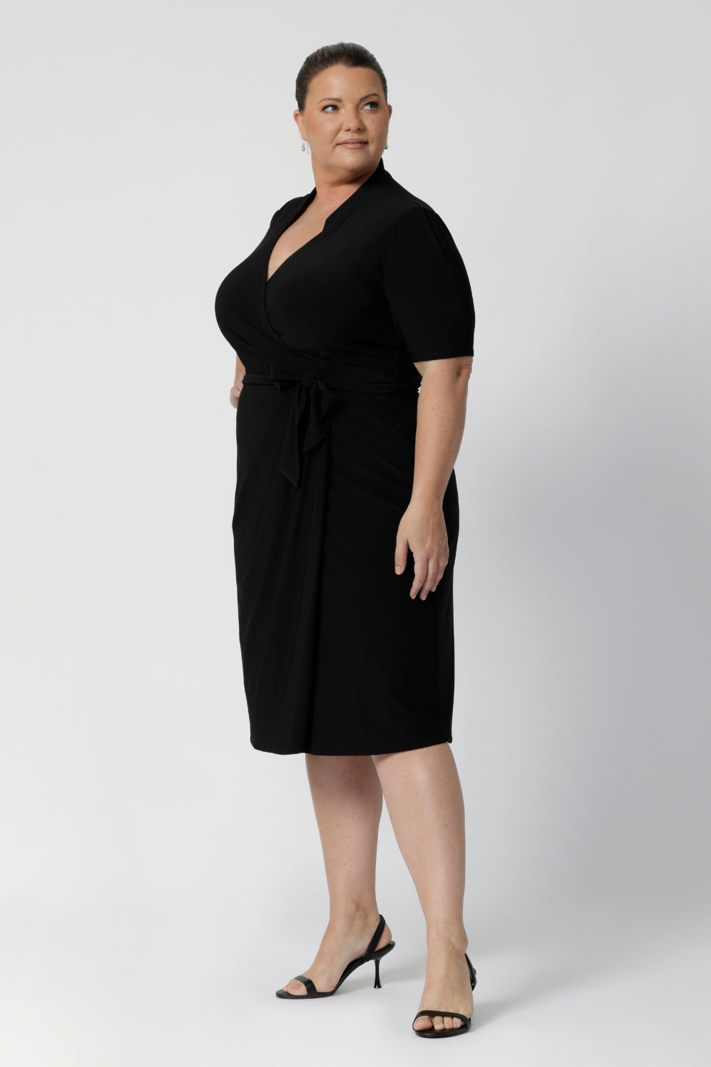 A size 18 woman wears the Kris dress in black. A functioning wrap work dress in soft black jersey. Great for being comfortable in the office. Made in Australia for women size inclusive fashion 8 - 24.