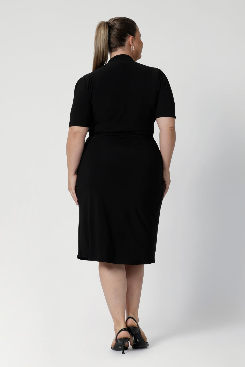 Back view of a size 18 woman wears the Kris dress in black. A functioning wrap work dress in soft black jersey. Great for being comfortable in the office. Made in Australia for women size inclusive fashion 8 - 24.