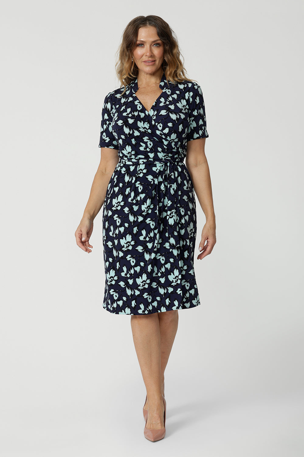 Showing a good wrap dress for work and event wear, a size 12 over 40 woman wears a short sleeve wrap dress in blue floral print. This easy care, jersey wrap dress is made in Australia by Australian and New Zealand women's clothing brand, Leina & Fleur.