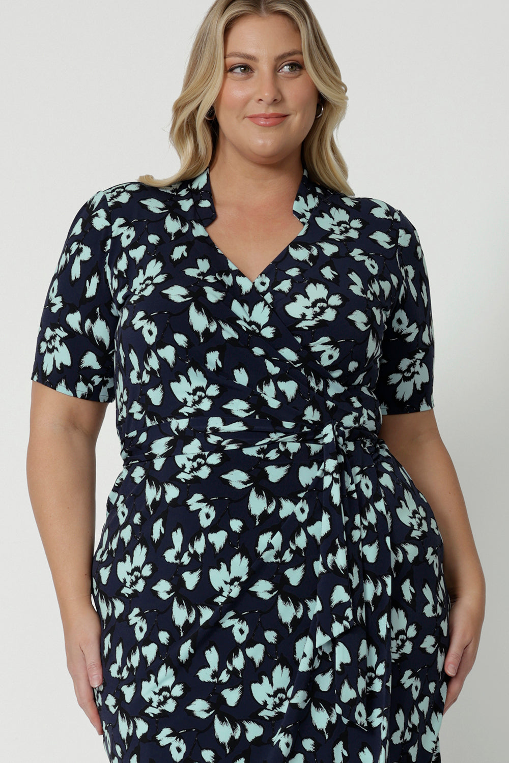 Showing the collar and wrap detail of a good wrap dress for plus size women, a size 18, curvy woman wears a short sleeve wrap dress in blue floral print. This easy care, jersey wrap dress is made in Australia by Australian and New Zealand women's clothing brand, Leina & Fleur.