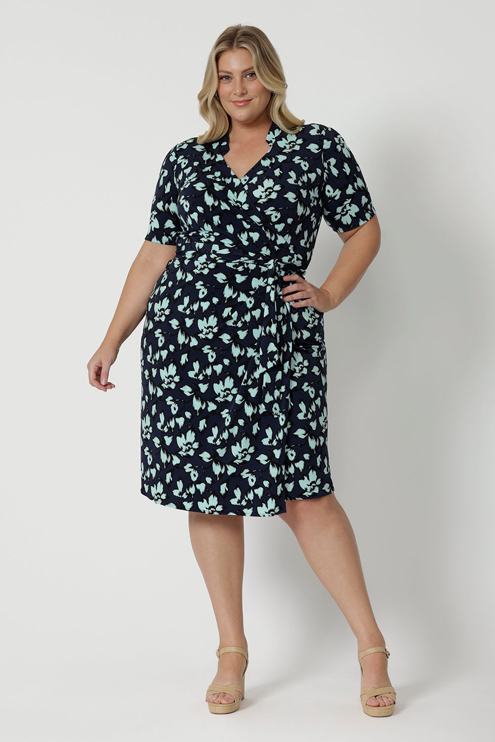 Showing a good wrap dress for plus size women, a size 18, curvy woman wears a short sleeve wrap dress in blue floral print. This easy care, jersey wrap dress is made in Australia by Australian and New Zealand women's clothing brand, Leina & Fleur.