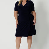 Full body image of plus sized model wears navy wrap dress. Light weight dress is made of jersey fabric. Australian workwear dress made for woman in Australia and New Zealand. Leina & Fleur is a woman’s clothing brand that stocks sizes from 8 to 24 including plus sizes and petite sizes. Shop this look - Kris Dress in Navy.