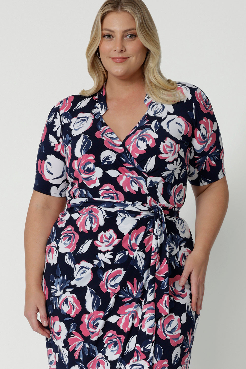 Close up shot of plus sized woman wearing floral wrap dress. The dress design made from easy care dry touch jersey fabric. Printed design has a colourful combination of white and pink roses on navy base. This work wear dress is comfortable, easy and perfect for any travel capsule. Designed in Australia by Australian and New Zealand women. Leina & Fleur is a fashion brand for woman that have sizes from 8 to 24. Shop this functioning wrap dress in petite sizes and plus sizes.