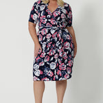 Full body front view of plus sized woman wearing a printed floral dress. This wrap dress has a floral design made from dry touch jersey fabric and has a colourful combination of white and pink roses on navy base. This work wear dress is easy and comfortable, perfect for travel. Easy care fabric used to make wrap dress in Australia for New Zealand and Australian women. Leina & Fleur is a clothing company for woman that stock sizes from 8 to 24. Shop this functioning wrap dress in plus sizes and petite sizes.