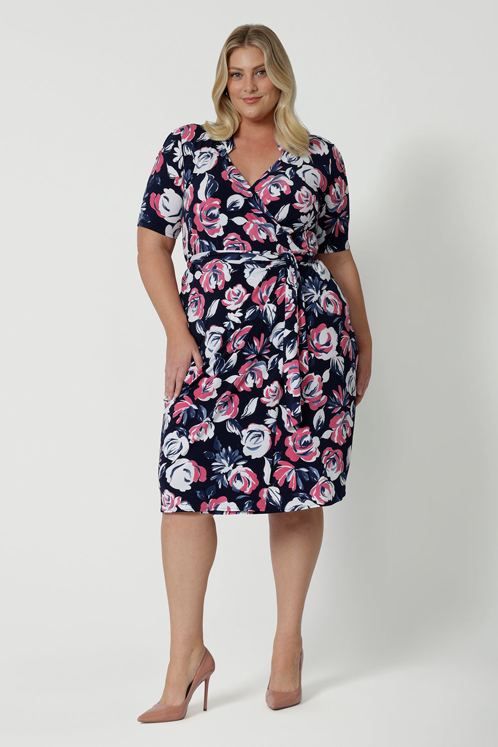 Full body front view of plus sized woman wearing a printed floral dress. This wrap dress has a floral design made from dry touch jersey fabric and has a colourful combination of white and pink roses on navy base. This work wear dress is easy and comfortable, perfect for travel. Easy care fabric used to make wrap dress in Australia for New Zealand and Australian women. Leina & Fleur is a clothing company for woman that stock sizes from 8 to 24. Shop this functioning wrap dress in plus sizes and petite sizes.