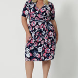 Full body front view of plus sized woman wearing a printed floral dress. This wrap dress has a floral design made from dry touch jersey fabric and has a colourful combination of white and pink roses on navy base. This work wear dress is easy and comfortable, perfect for travel. Easy care fabric used to make wrap dress in Australia for New Zealand and Australian women. Leina & Fleur is a clothing company for woman that stock sizes from 8 to 24. Shop this functioning wrap dress in plus sizes and petite sizes.