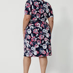 Back shot image of plus sized woman wearing a floral wrap dress. Dress is made from easy care dry touch jersey fabric. Printed design displays white and pink roses on navy. This work wear dress is comfortable, easy and perfect for travel. Outfit designed in Australia by Australian and New Zealand women. Leina and Fleur is a fashion brand for woman that stock sizes from 8 to 24. Shop this functioning wrap dress in petite sizes and plus sizes.