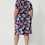 Back shot image of plus sized woman wearing a floral wrap dress. Dress is made from easy care dry touch jersey fabric. Printed design displays white and pink roses on navy. This work wear dress is comfortable, easy and perfect for travel. Outfit designed in Australia by Australian and New Zealand women. Leina and Fleur is a fashion brand for woman that stock sizes from 8 to 24. Shop this functioning wrap dress in petite sizes and plus sizes.