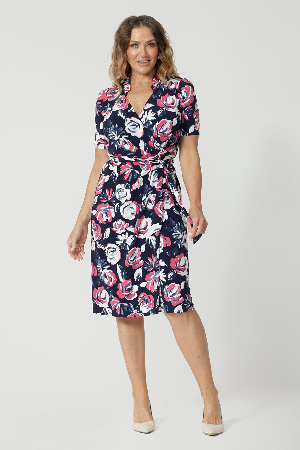 Full body front view of 40 year old woman wearing short sleeved dress. This wrap dress displays a floral design. Made of easy care dry touch jersey fabric. Colourful floral dress has a combination of pink and white roses on navy dress. This work wear dress is comfortable and perfect for travel. Made for Australian and New Zealand women. Leina & Fleur is a women's clothing brand that proudly stocks sizes from 8 to 24. Shop this functioning wrap dress in plus sizes and petite sizes.