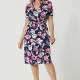 Full body front view of 40 year old woman wearing short sleeved dress. This wrap dress displays a floral design. Made of easy care dry touch jersey fabric. Colourful floral dress has a combination of pink and white roses on navy dress. This work wear dress is comfortable and perfect for travel. Made for Australian and New Zealand women. Leina & Fleur is a women's clothing brand that proudly stocks sizes from 8 to 24. Shop this functioning wrap dress in plus sizes and petite sizes.