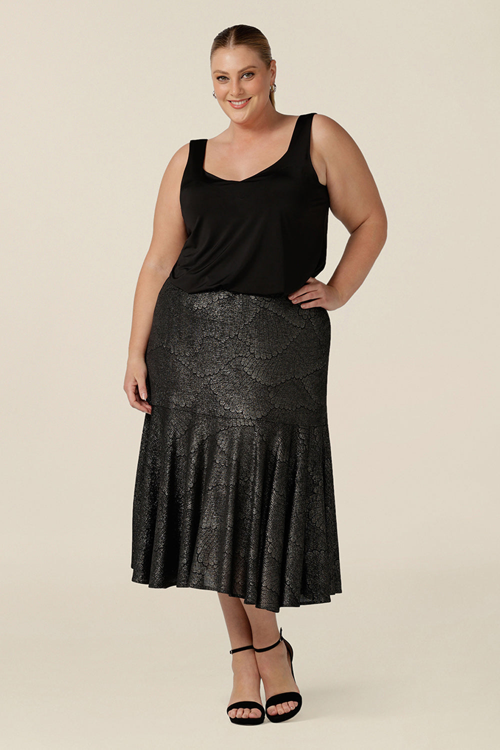 A size 18, plus size woman wears a midi length, foil print evening skirt with ruffle hem. For cocktail and evening wear, this elegant skirt is worn with a black cami top. Both cocktail skirt and top are made in Australia by women's fashion brand, Leina & Fleur. Shop their evening and cocktail wear online in petite to plus sizes.