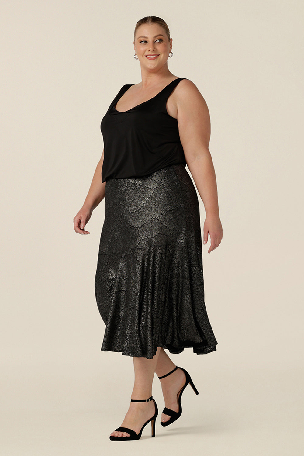 Side view of a size 18, plus size woman wears a midi length, foil print evening skirt with ruffle hem. For cocktail and evening wear, this elegant skirt is worn with a black cami top. Both cocktail skirt and top are made in Australia by women's fashion brand, Leina & Fleur. Shop their evening and cocktail wear online in petite to plus sizes.