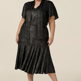 A plus size, size 18 woman wears a good top for curvy women's evening and event wear. In shimmering bronze foil print, this V-neck top with short flutter sleeves is worn with midi length foil print skirt with ruffle hem. Styled together as a faux cocktail dress, both going out top and skirt are made in Australia by women's occasionwear brand, Leina and Fleur.