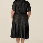 Back view of a plus size, size 18 woman wearing a good top for curvy women's evening and event wear. In shimmering bronze foil print, this V-neck top with short flutter sleeves is worn with midi length foil print skirt with ruffle hem. Styled together as a faux cocktail dress, both going out top and skirt are made in Australia by women's occasionwear brand, Leina and Fleur.