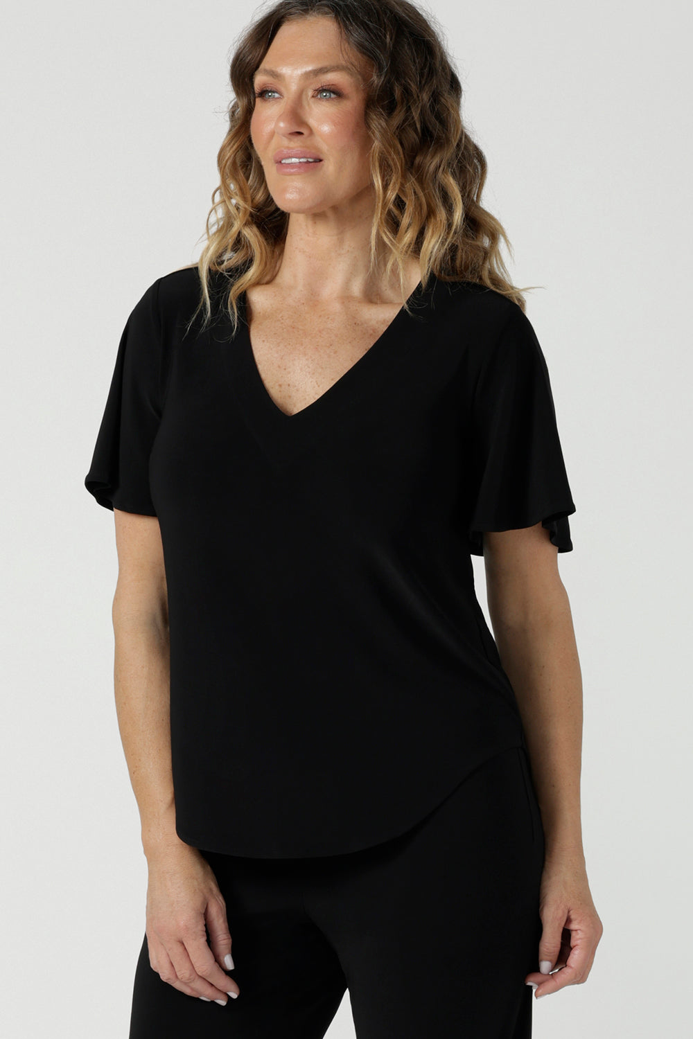 Close up of a size 10 woman wears the Lila Top in Black, a jersey workwear top for women for flutter sleeve and v-neckline. Made in Australia for women size 8-24. Styled back with black Paxton pants.