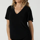 Close up of a size 10 woman wears the Lila Top in Black, a jersey workwear top for women for flutter sleeve and v-neckline. Made in Australia for women size 8-24. Styled back with black Paxton pants.