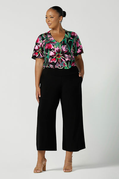 A plus size, size 18 woman wears a floral print jersey, V-neck top with flutter sleeves with wide leg black pants. A good top for summer casual wear, or style tucked as a workwear top. Shop made in Australia tops in petite to plus sizes online at Australian fashion brand, Leina & Fleur.