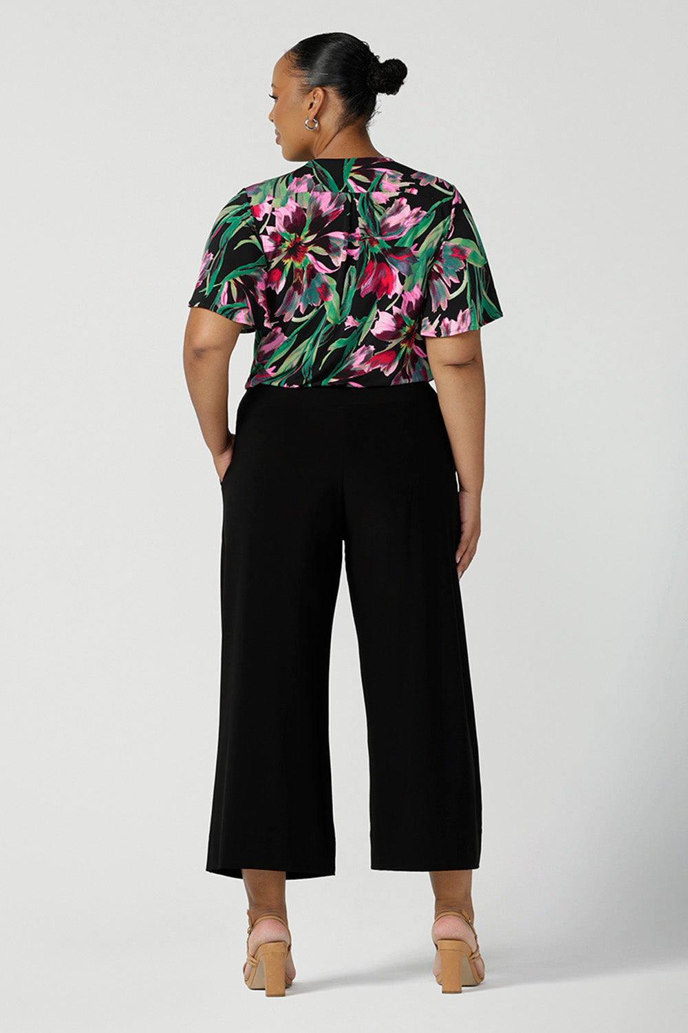 Back view of a plus size, size 18 woman wearing a floral print jersey, V-neck top with flutter sleeves with wide leg black pants. A good top for summer casual wear, or style tucked as a workwear top. Shop made in Australia tops in petite to plus sizes online at Australian fashion brand, Leina & Fleur.