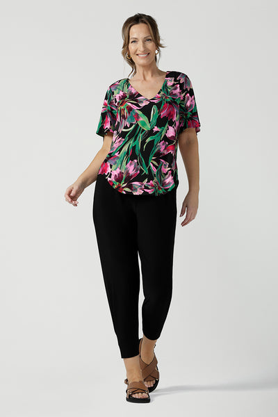 A size 10, 40 plus woman wears a floral print jersey, V-neck top with flutter sleeves with tapered leg black travel pants. A good top for summer casual wear, or style tucked as a workwear top. Shop made in Australia tops in petite to plus sizes online at Australian fashion brand, Leina & Fleur.