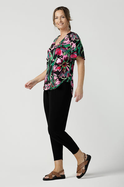 Side view of a size 10, 40 plus woman wearing a floral print jersey, V-neck top with flutter sleeves with tapered leg black travel pants. A good top for summer casual wear, or style tucked as a workwear top. Shop made in Australia tops in petite to plus sizes online at Australian fashion brand, Leina & Fleur.