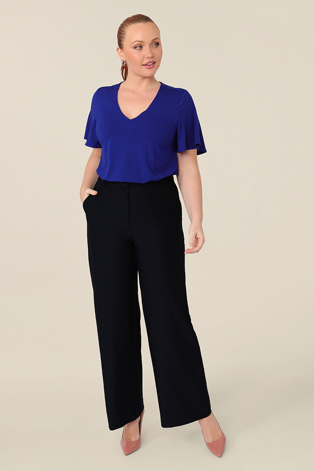 A size 12 curvy woman wears a blue bamboo jersey top with a V-neck and short flutter sleeves. It is a lightweight and breathable top thanks to its natural and sustainable bamboo fibres, and being made in Australia, it is part of L&Fs move towards greater sustainability and eco-conscious fashion. Styled with a navy wide leg pant, perfect for work wear. 