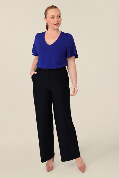 A size 12 curvy woman wears a blue bamboo jersey top with a V-neck and short flutter sleeves. It is a lightweight and breathable top thanks to its natural and sustainable bamboo fibres, and being made in Australia, it is part of L&Fs move towards greater sustainability and eco-conscious fashion. Styled with a navy wide leg pant, perfect for work wear. 