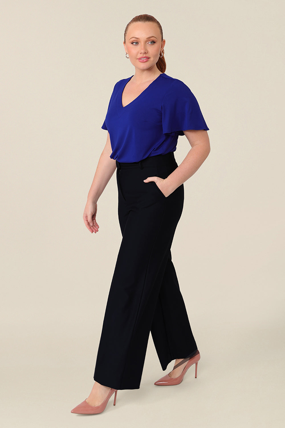 A size 12 curvy woman wears a blue bamboo jersey top with a V-neck and short flutter sleeves. It is a lightweight and breathable top thanks to its natural and sustainable bamboo fibres, and being made in Australia, it is part of L&Fs move towards greater sustainability and eco-conscious fashion. Styled with a navy wide leg pant, perfect for work wear.