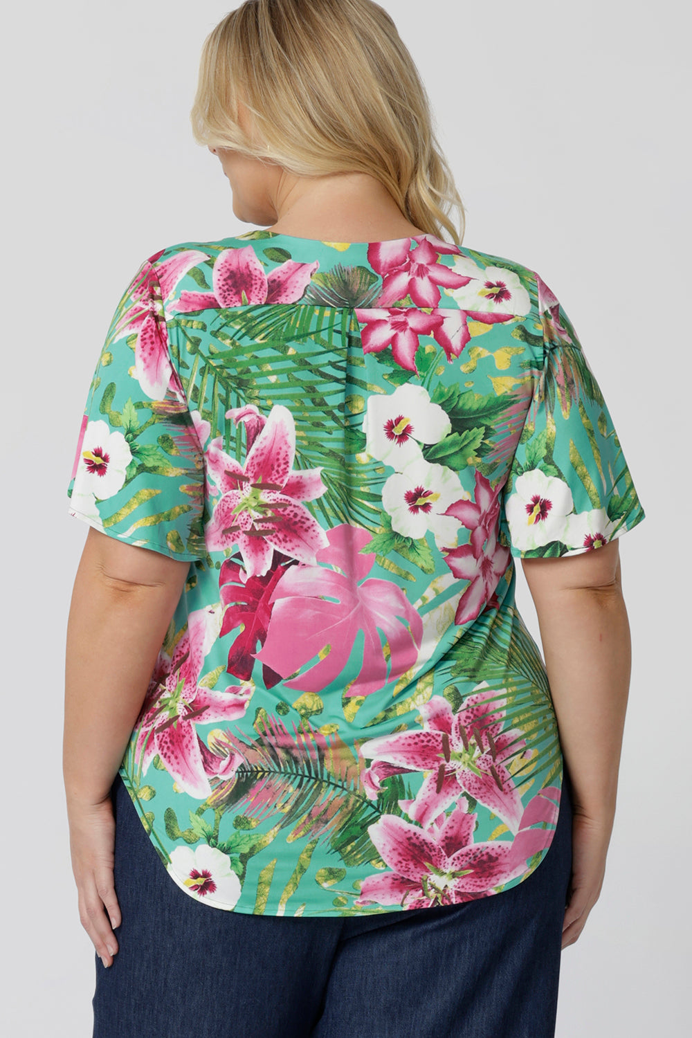 Back view of floral top, featuring shirt tail hemline and flutter sleeves. The Malibu print is on a teal-blue slinky jersey base with print details in shades of green, pink, white, yellow and brown. The fabric has a soft stretch and cooling effect. Sizes 8-24 are now available to shop.