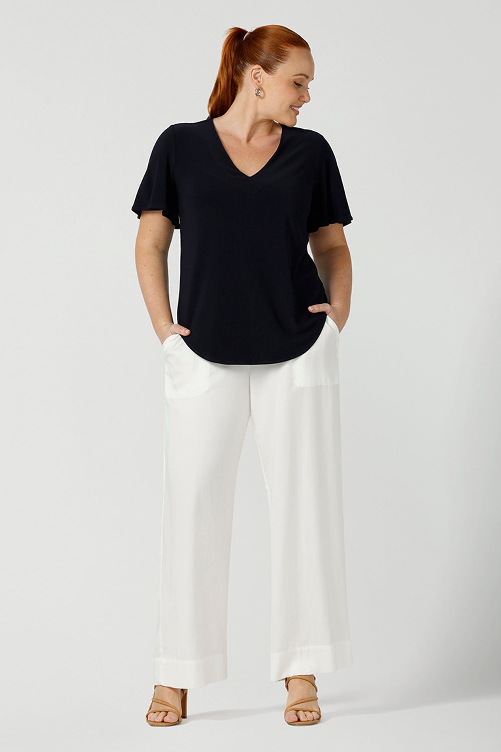 Worn by a size 12, curvy woman, this is a semi-fitted, V-neck top with flutter sleeves in navy blue jersey. Made in Australia by women's clothing brand, Leina & Fleur , this classic navy top is worn with wide leg white pants for summer.  Great for capsule wardrobes, smart casual wear and as a work blouse, shop this top and other Australian-made women's clothing online in sizes 8 to 24 at Leina & Fleur's online fashion boutique!