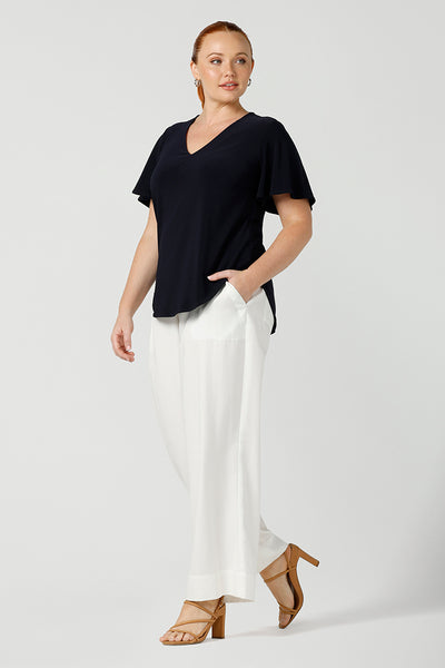 Worn by a size 12, curvy woman, this is a semi-fitted, V-neck top with flutter sleeves in navy blue jersey. Made in Australia by ladies clothing brand, Leina & Fleur , this classic navy top is worn with wide leg white pants for summer. Great for capsule wardrobes, smart casual wear and as a work blouse, shop this top and other Australian-made women's clothing online in an inclusive size range of sizes 8 to 24 at Leina & Fleur's online boutique!