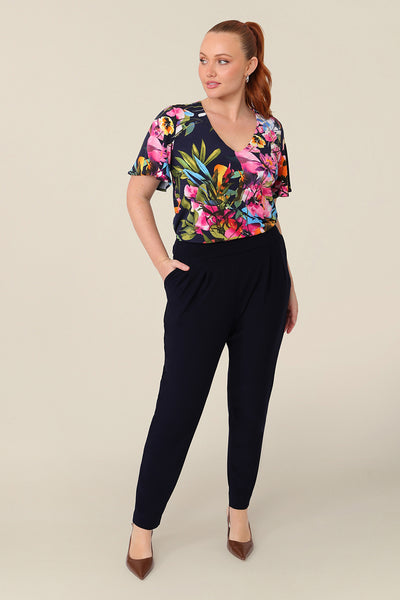 A size 12 curvy woman wears a floral jersey top with a V-neck and short flutter sleeves. It is a lightweight and crease-free top thanks to its stretch jersey fabric. Styled with a tapered leg navy trouser for an everyday look. Made in Australia, for sizes 8-24.