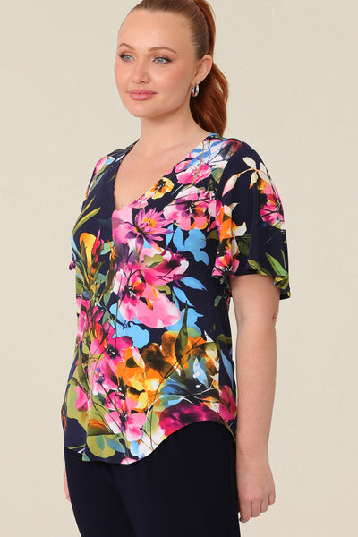 A size 12 curvy woman wears a floral jersey top with a V-neck and short flutter sleeves. It is a lightweight and crease-free top thanks to its stretch jersey fabric. Made in Australia, for sizes 8-24.