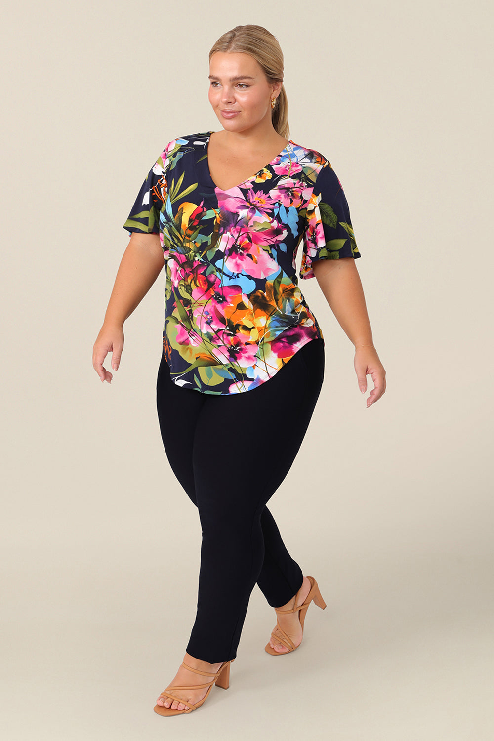 A plus size, size 14 woman wears a V-neck floral top with short flutter sleeves. Made from dry-touch jersey, and styled with a fitted navy trouser,this top is comfortable for everyday style. Designed and made in Australia for petite to plus size women.