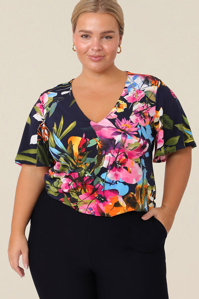 A plus size, size 14 woman wears a V-neck floral top with short flutter sleeves. Made from dry-touch jersey, this top is comfortable for everyday style. Designed and made in Australia for petite to plus size women.