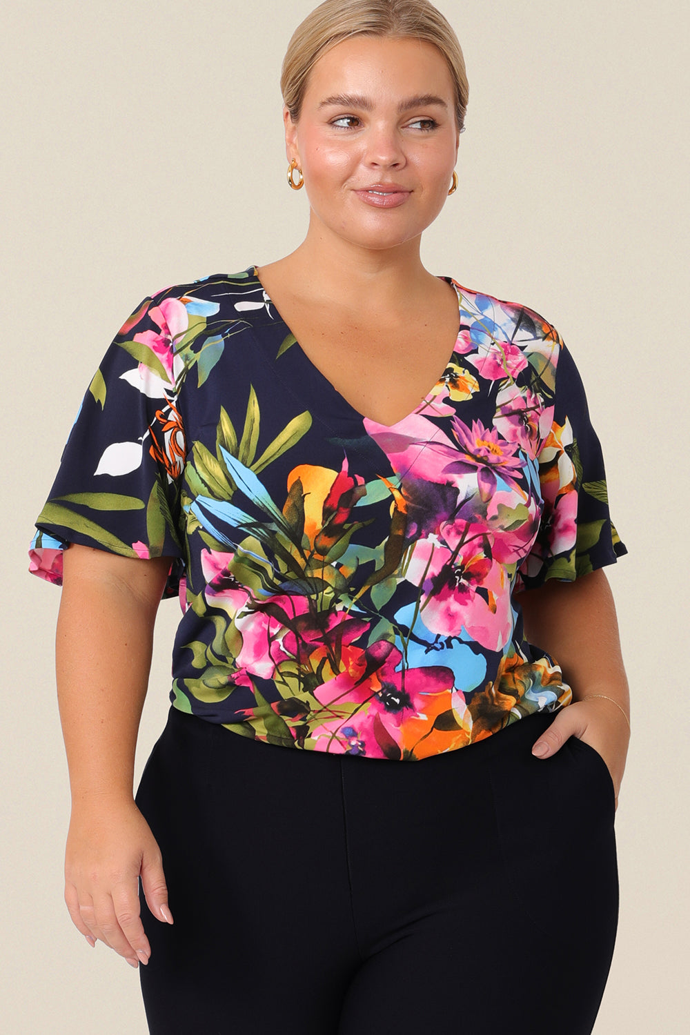 A plus size, size 14 woman wears a V-neck floral top with short flutter sleeves. Made from dry-touch jersey, this top is comfortable for everyday style. Designed and made in Australia for petite to plus size women.